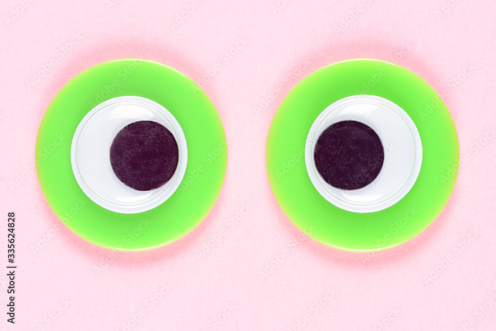 Googly toy eyes. Neon green mad cross-eyed eyes look straight at you, on a background of rose paper. Macro close-up. Googly toy eyes. Neon green mad cross-eyed eyes look straight at you, on a background of rose paper. Macro close-up.