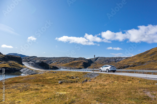 White car driving Norway highway mountain road on sunny autumn bright day. Wild nature travel, blue sky, electric car, eco life