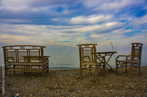 Empty wooden chairs and tables lined up on pebble beach. Seaside restaurant ready for sunny weekend customers. Relaxing place for evening or night meal. Outdoor resort cafe for eating or drinking.