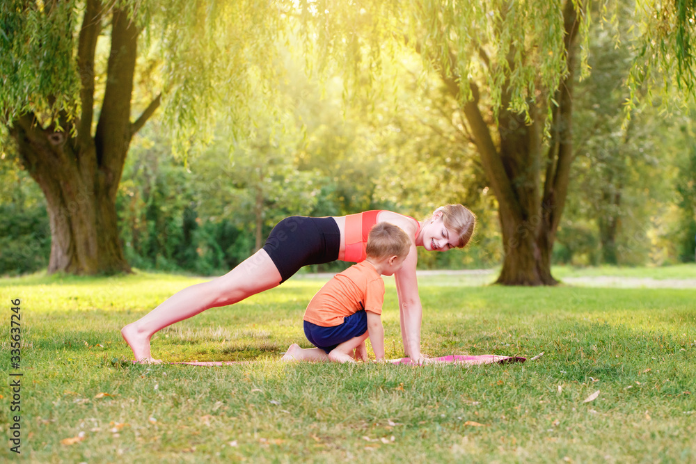 Fototapeta premium Family summer outdoor sport activity. Young Caucasian mother with child toddler boy doing workout yoga fitness in park. Woman mom doing physical exercises together with a little kid outside.