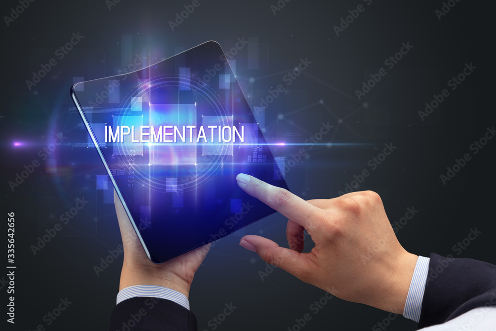 Businessman holding a foldable smartphone with IMPLEMENTATION inscription, new technology concept Businessman holding a foldable smartphone with IMPLEMENTATION inscription, new technology concept