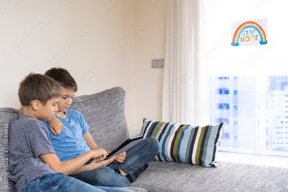 Online learning at home, distance learning during Coronavirus quarantine. Boy with tablet computer sitting in the room. On the window drawing picture with inscription Stay at home Online learning at home, distance learning during Coronavirus quarantine. Boy with tablet computer sitting in the room. On the window drawing picture with inscription Stay at home