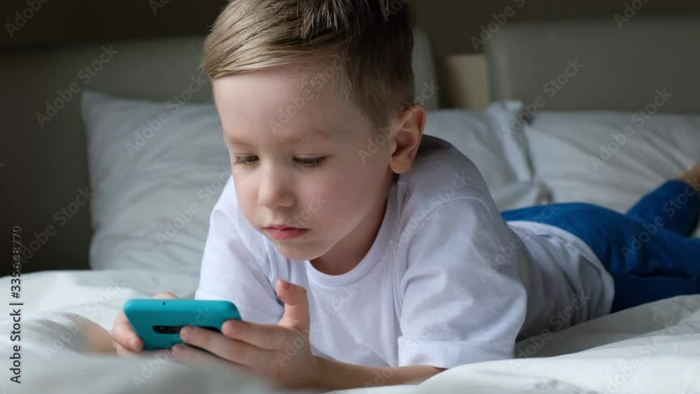 Cute little boy toddler playing mobile game, lying on a bed and holding smartphone Cute little boy toddler playing mobile game, lying on a bed and holding smartphone