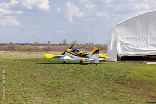 Sports aerobatics, painted in white and yellow , is parked with grass on an open airfield awaiting departure.