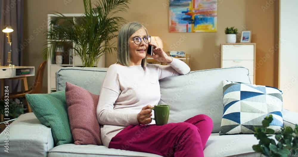 Happy Caucasian pretty old woman with cup of coffee talking on phone while sitting on sofa in cozy room. Beautiful joyful senior female with hot drink calling on cellphone indoors. Home concept Happy Caucasian pretty old woman with cup of coffee talking on phone while sitting on sofa in cozy room. Beautiful joyful senior female with hot drink calling on cellphone indoors. Home concept