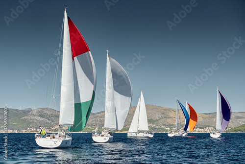 Sailboats compete in a sail regatta at sunset, race of sailboats, reflection of sails on water, multicolored spinnakers, number of boat is on aft boats, island is on background, clear weather
