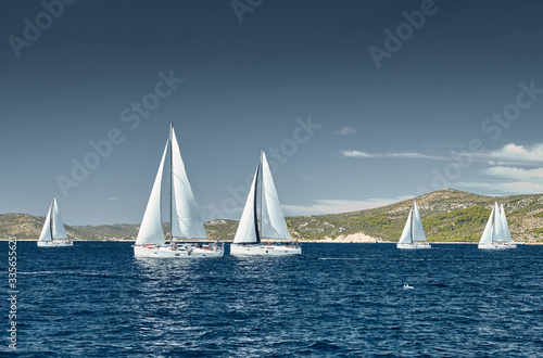 Fototapeta Naklejka Na Ścianę i Meble -  Sailboats compete in a sail regatta at sunset, race of sailboats, reflection of sails on water, multicolored spinnakers, number of boat is on aft boats, island is on background, clear weather