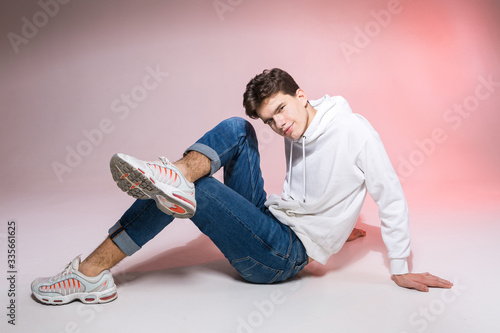 Handsome young stylish man sitting on the floor in studio. Teenager model posing while sitting on a pink background in casual clothes. The guy in jeans, sneakers and a white jacket with a hood