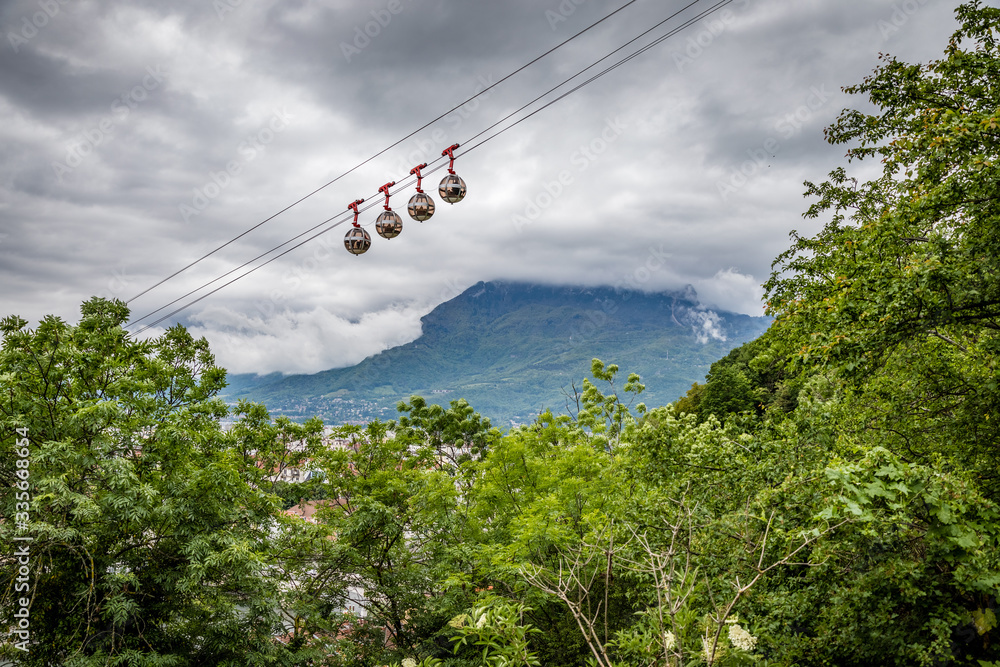 Set of 4 gondola pods heading up the mountain with dark clouds in the background and some distant green mountains in Grenoble France