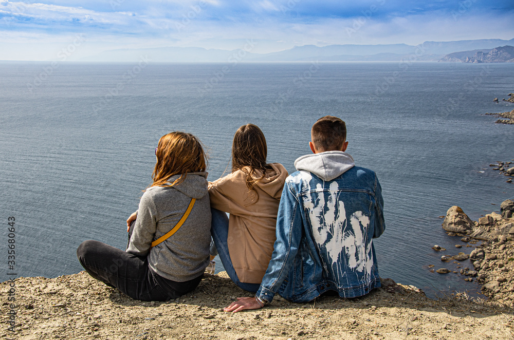 Fototapeta premium A guy and two girls are sitting on a mountain and looking at the sea. Travel and adventure, tourism.