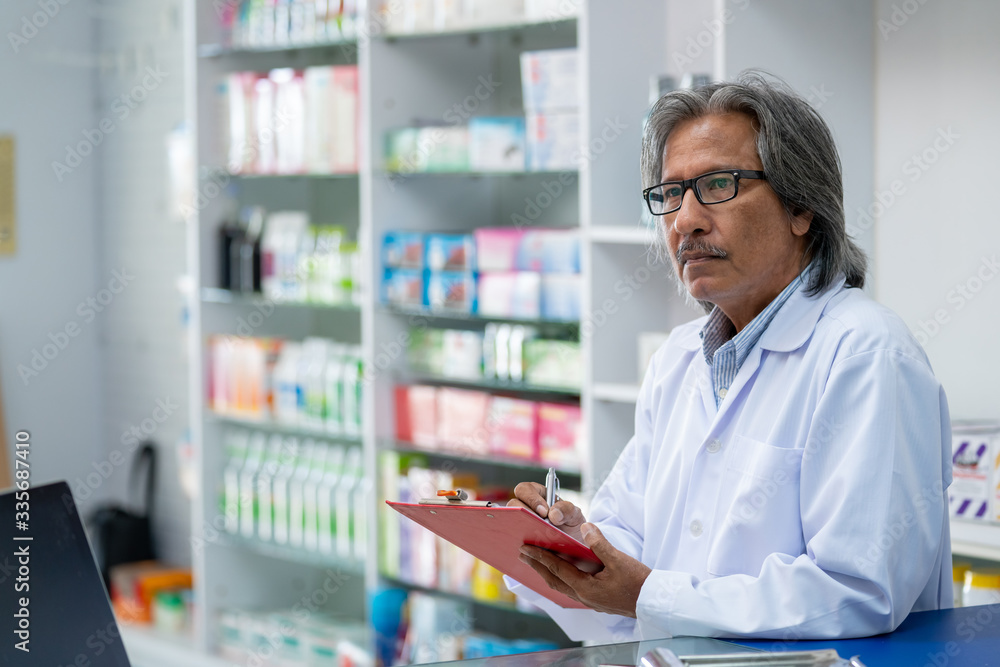 Professional senior Asian male Pharmacist checking medicines and health product inventory stock at hospital pharmacy or drugstore. Medical, pharmaceutics and personal healthcare concept. Professional senior Asian male Pharmacist checking medicines and health product inventory stock at hospital pharmacy or drugstore. Medical, pharmaceutics and personal healthcare concept.