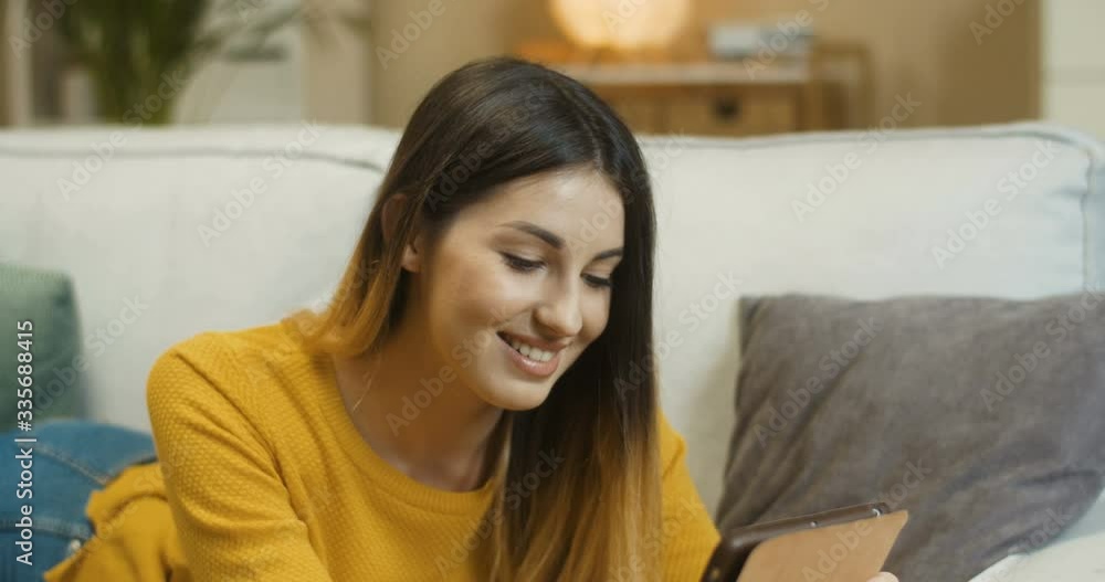 Joyful pretty Caucasian young female searching on device at home in quarantine. Close up portrait of happy smiling beautiful girl typing on tablet while lying on sofa in living room. Rest concept Joyful pretty Caucasian young female searching on device at home in quarantine. Close up portrait of happy smiling beautiful girl typing on tablet while lying on sofa in living room. Rest concept