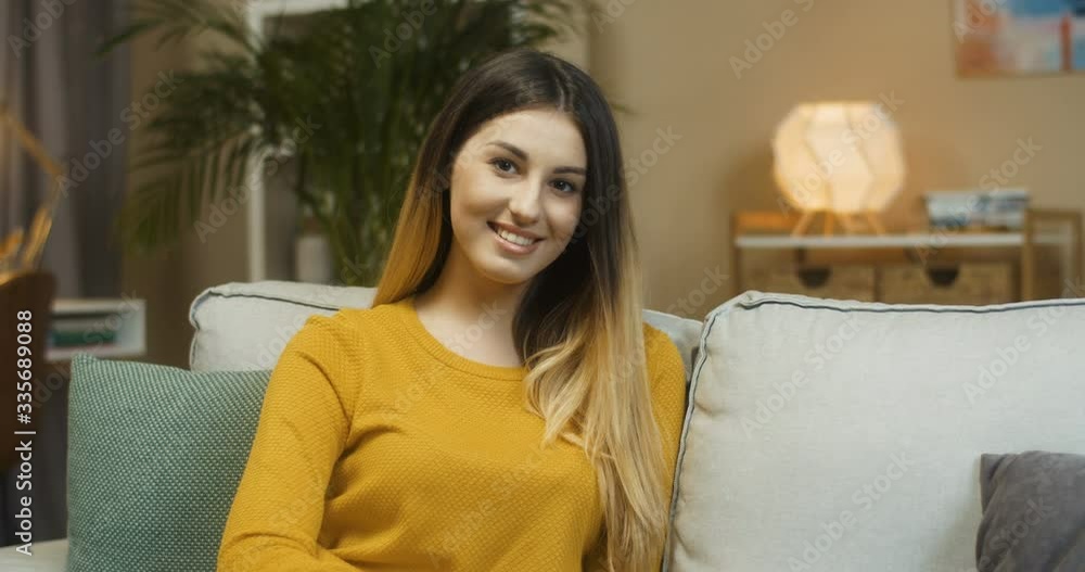 Cheerful Caucasian woman smiling to camera while sitting on couch indoor. Close up portrait of pretty happy young female model posing with smile on face in cozy living room at home. Isolation concept Cheerful Caucasian woman smiling to camera while sitting on couch indoor. Close up portrait of pretty happy young female model posing with smile on face in cozy living room at home. Isolation concept