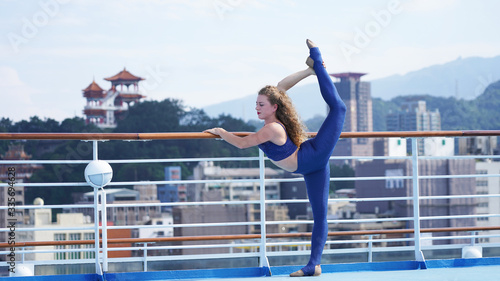 Beautiful slim European girl curly hair on observation deck of ship, against the background of Asian city. Blond gymnast standing on one leg in split on railing, sunny day.Professional circus acrobat