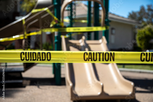 Yellow caution tape in english and spanish barring access to playgound equipment at a park. The access is denied due to the Coronavirus Pandemic