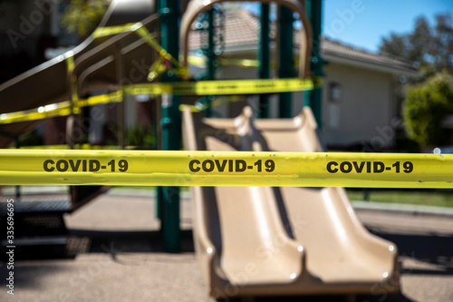 Yellow caution tape stating COVID-19 denying access to playgound equipment at a park. The access is denied due to the Coronavirus Pandemic
