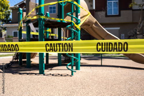 Yellow caution tape in english and spanish barring access to playgound equipment at a park. The access is denied due to the Coronavirus Pandemic