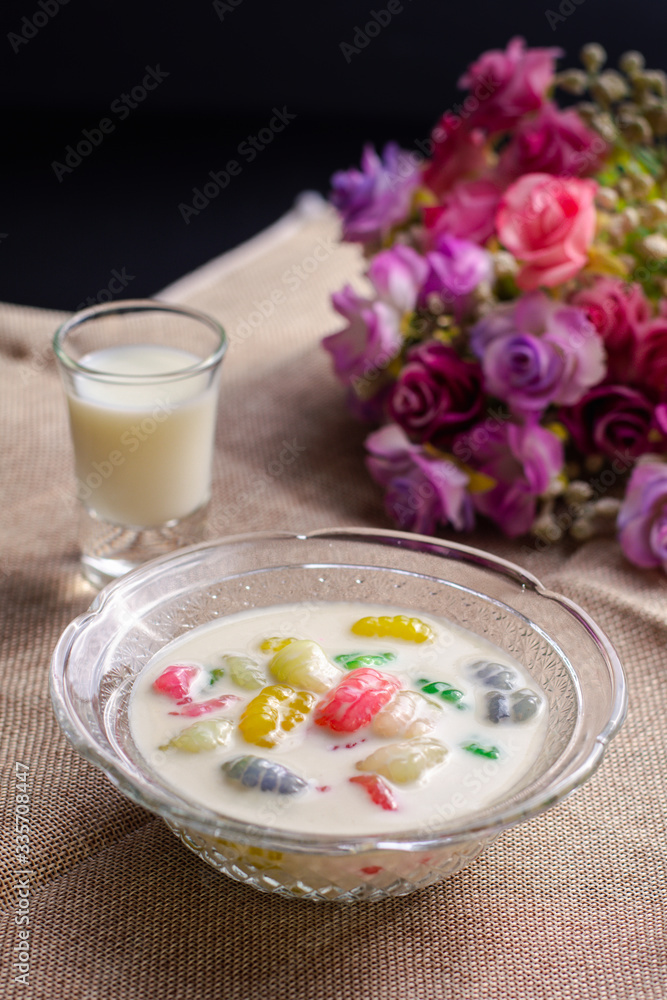Colorful sweet rice gnocchi in fresh coconut milk in glass bowl on brown tablecloth on blackground. Delicious Thai desserts.