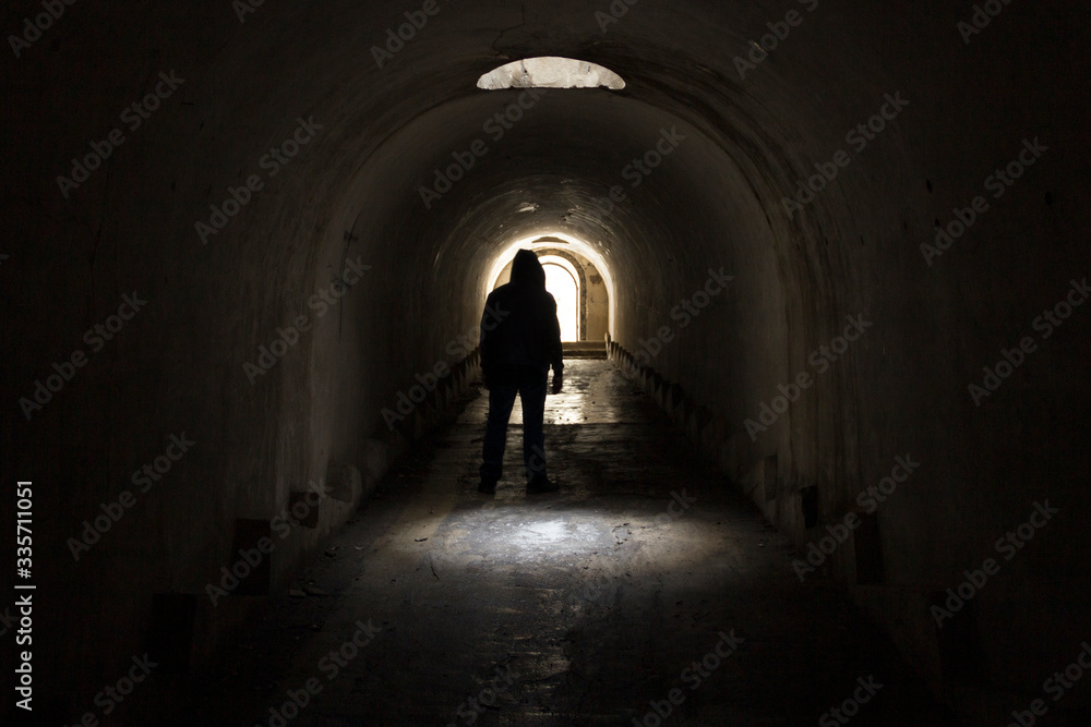 A man in dark clothes with a hood going to the exit of the light from a dark gloomy underground tunnel, lit by solar light from a hatch in the ceiling of the corridor.
