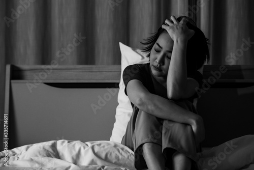 Monochrome Depressed young beautiful Asian woman sitting alone on bed at night. Stressed woman holding head in hand think of the problem seriously. Mental health, healthcare and social issue concept.