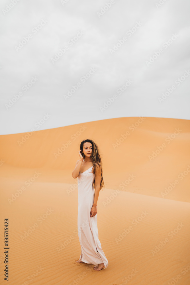 Pretty portrait of young beautiful woman in sand dunes of moroccan Sahara desert. Brunette with long hair, eastern appearance. Bride in white silk dress.