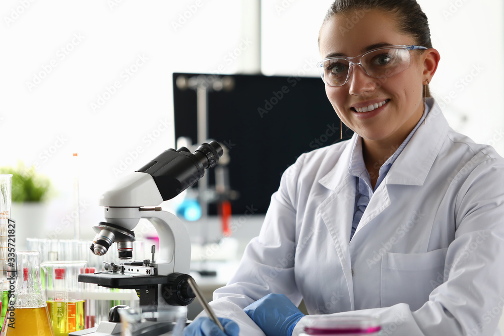 Portrait of smiling woman looking at camera with happiness. Joyful laboratory assistant working with chemicals in lab. Flasks, test-tubes, beakers, microscope on table. Chemical experiments concept Portrait of smiling woman looking at camera with happiness. Joyful laboratory assistant working with chemicals in lab. Flasks, test-tubes, beakers, microscope on table. Chemical experiments concept