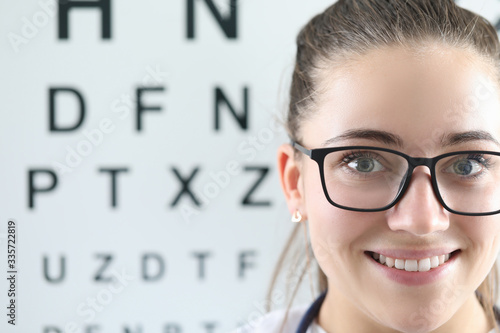 Close-up of cheerful female ophthalmologist smiling on camera. Special board with letters to checkup vision. Copy space in left side. Modern medicine healthcare and eye doctor concept