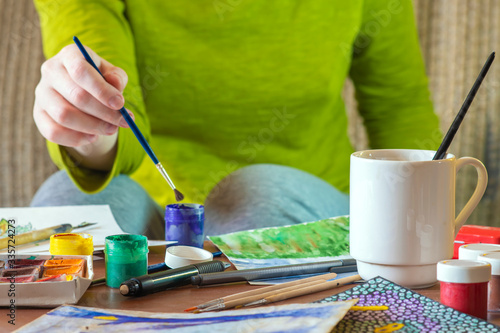 A girl whose face is not visible draws at the table covered with drawing accessories - paper, brushes, paint cans and others.