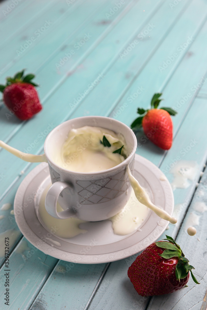 Red ripe strawberries and a white coffee Cup, on a turquoise wooden table. The berry falls into the milk. Frozen movement. Splashes from the Cup. Red ripe strawberries and a white coffee Cup, on a turquoise wooden table. The berry falls into the milk. Frozen movement. Splashes from the Cup.