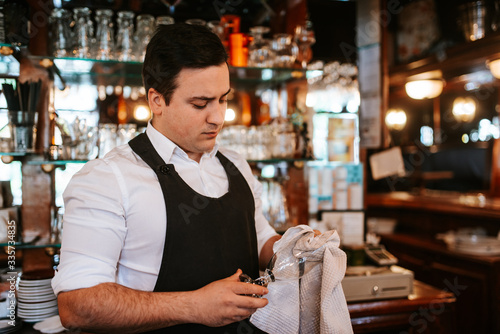 A young caucasian waiter polishes glasses at a bar