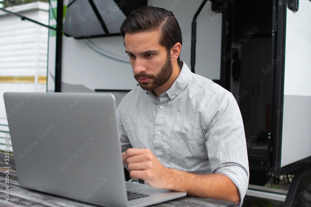 A man wearing a long sleeve grey button down business shirt with brown slicked comb over hair working on laptop on a picnic table remotely near his camper able to avoid the busy society work community A man wearing a long sleeve grey button down business shirt with brown slicked comb over hair working on laptop on a picnic table remotely near his camper able to avoid the busy society work community