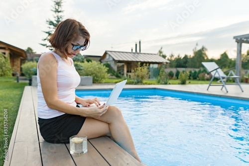 Mature woman with laptop near private home pool, successful beautiful female freelancer