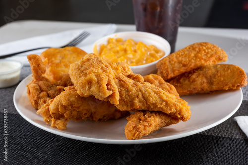A view of a plate of breaded chicken tenders, with potato wedges and macaroni and cheese sides, in a restaurant or kitchen setting.