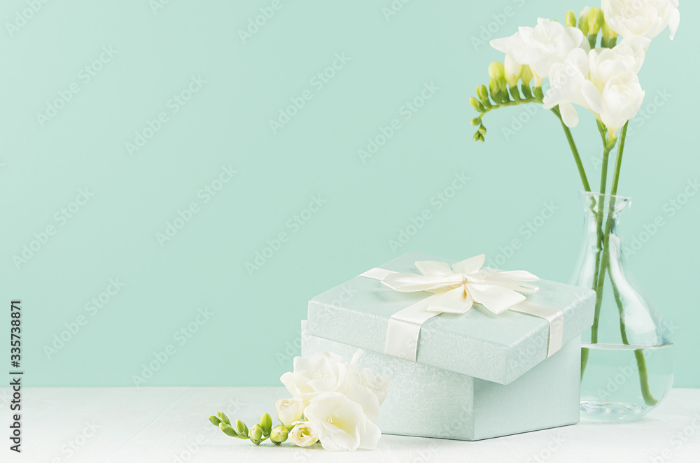 Fresh delicate white flowers freesia and festive ajar square gift box with ribbon and knot in green mint menthe interior on white wood board, closeup.