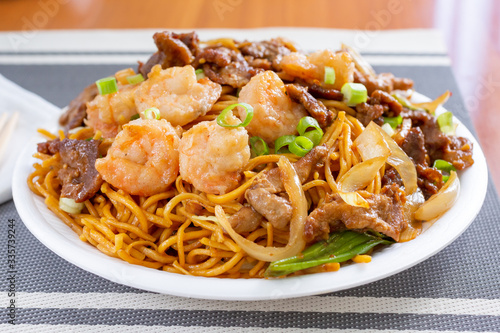 A view of a plate of Chinese chow mein featuring shrimp, beef and chicken, in a restaurant or kitchen setting.