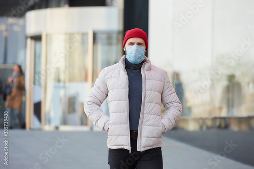  A man wearing a medical face mask to avoid the spread coronavirus (COVID-19). A guy with a mask on the face because of the pandemic holding hands in the pockets of the jacket in the center of the cit © Roman Tyukin