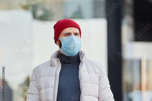 A man in a red watch cap wearing a medical face mask to avoid the spread coronavirus (COVID-19). A guy with a mask on the face because of the pandemic in the center of the city. Close-up portrait.