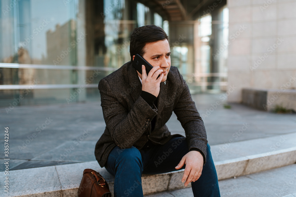 
A young businessman sits on a staircase in front of a building with a phone in his hand