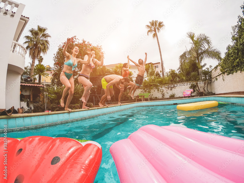 Group of happy friends jumping inside swimming pool at sunset - Young diverse culture people having fun in tropical vacation - Holiday, summer and friendship concept - Focus on guys bodies