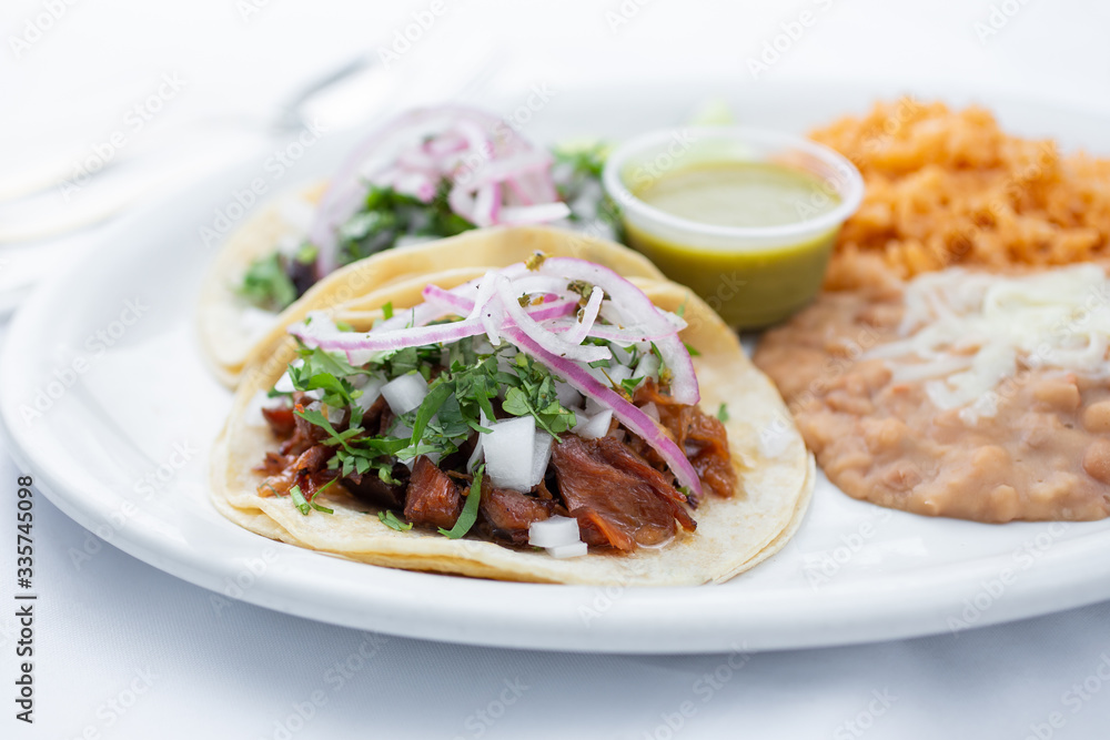 A view of two tacos al pastor with a side of beans and rice, in a restaurant or kitchen setting. A view of two tacos al pastor with a side of beans and rice, in a restaurant or kitchen setting.
