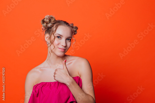 The girl in a red dress on a orange background in the studio. Blonde girl with two hair knots showing thumb up, looking to the camera and smiling.