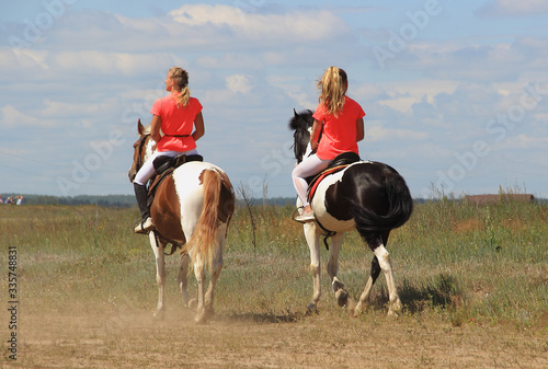 Two young horsewomen ride on piebald and black-and-white horses on a field against a cloudy sky