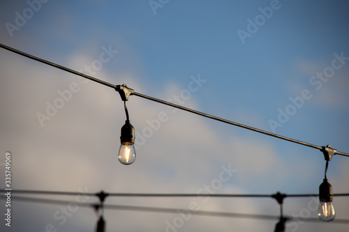 A string of outdoor lighting with on and off light bulbs hanging in the air, against a bright blue sky with clouds. Early morning feeling, selective focus on light bulbs.