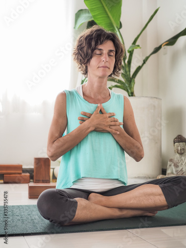 meditating woman with eyes closed and hands in heart mudra in crossed legged lotus position on a yoga mat in a studio with cushions buddha and palm tree