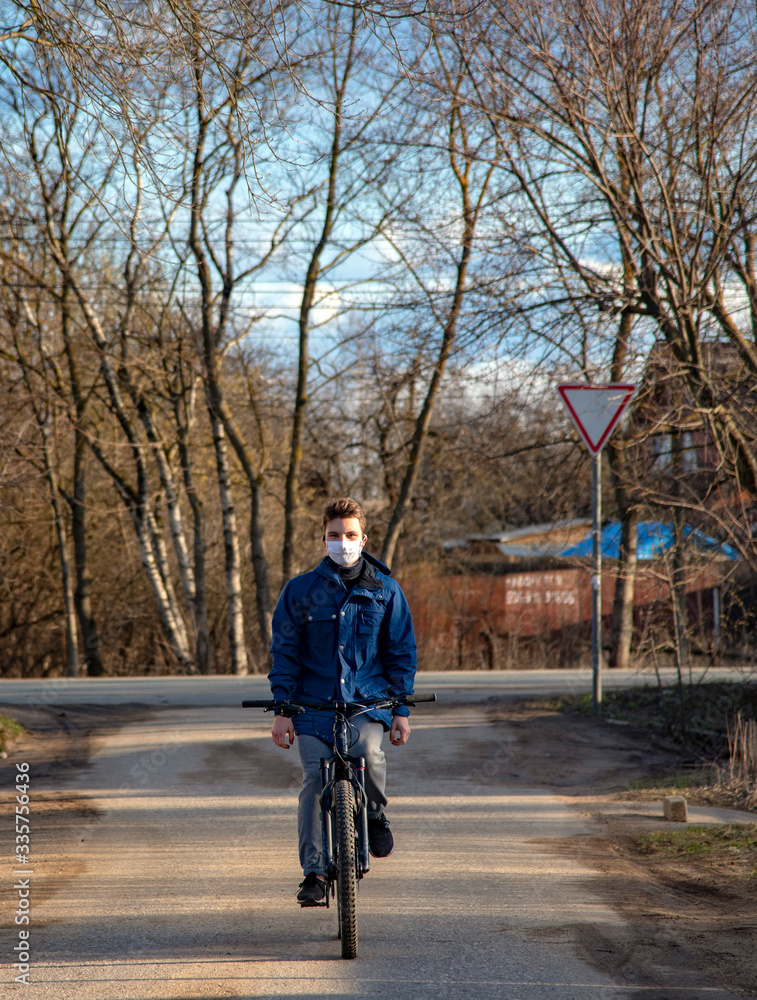 Obraz premium A young man in a protective medical mask and rubber gloves rides a Bicycle along a village street.