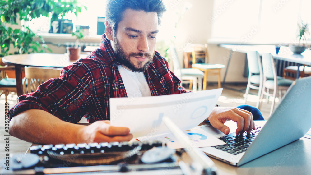 Handsome entrepreneur wearing casual clothes and stubble working in big bright room with laptop analyzing data, stats and graphs Handsome entrepreneur wearing casual clothes and stubble working in big bright room with laptop analyzing data, stats and graphs