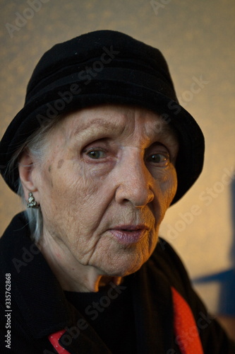 Portrait of an old woman. Grandmother in black stylish clothes and in a red scarf. Natural light from the window. Back light yellow. Face close up.