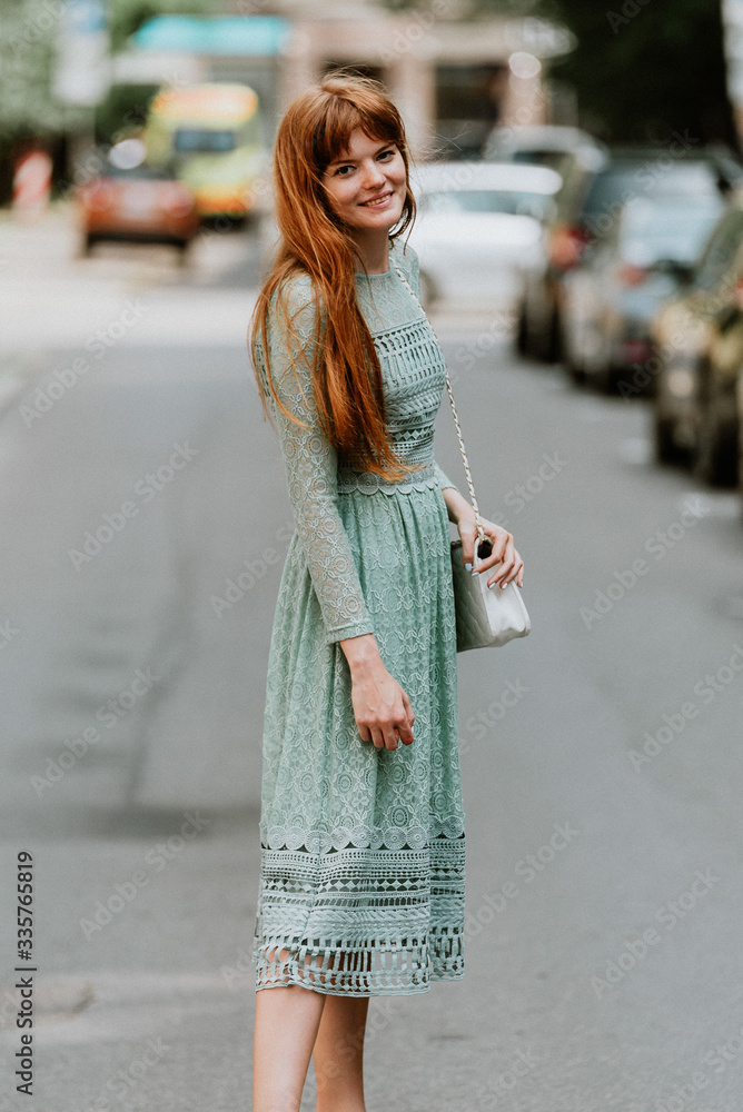 Stylish urban young redhair woman in a fashionable green dress posing in the street. European modern cute model girl enjoys a walk in the city. Spring fashion.