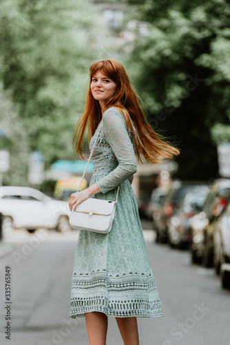 Stylish urban young redhair woman in a fashionable green dress posing in the street. European modern cute model girl enjoys a walk in the city. Spring fashion.