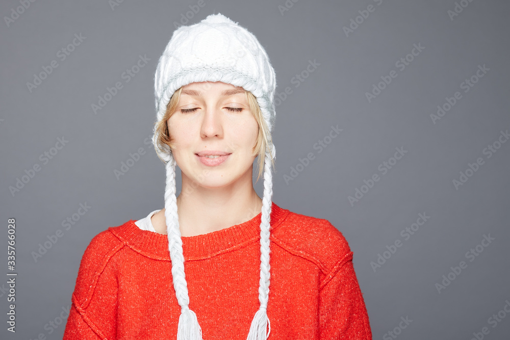 Portrait of gentle young blond short haired woman closes eyes, imagines pleasant plans, wears white hat, red winter sweater, poses over grey wall, copy space. Positive emotions, feelings concept.